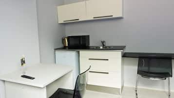 Una kitchenette compacta con encimera blanca, un microondas, una tetera y un lavabo. Hay un escritorio blanco pequeño con una silla y un televisor montado en la pared.