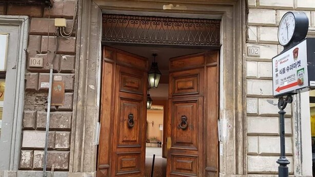 Una doble puerta de madera con herrajes de latón, una barandilla metálica arriba y una señal de calle a la derecha.