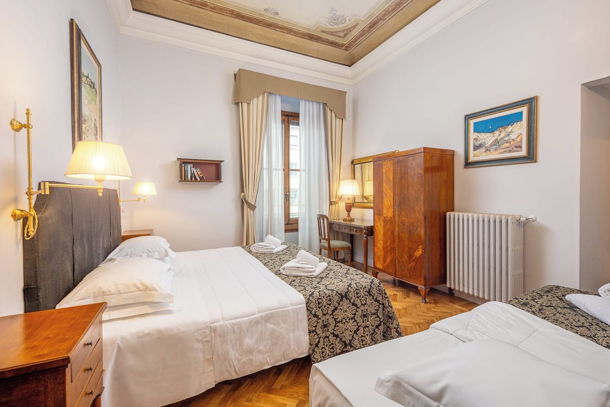 A hotel room with two beds, a desk, a wardrobe, a radiator, and a painting on the wall.