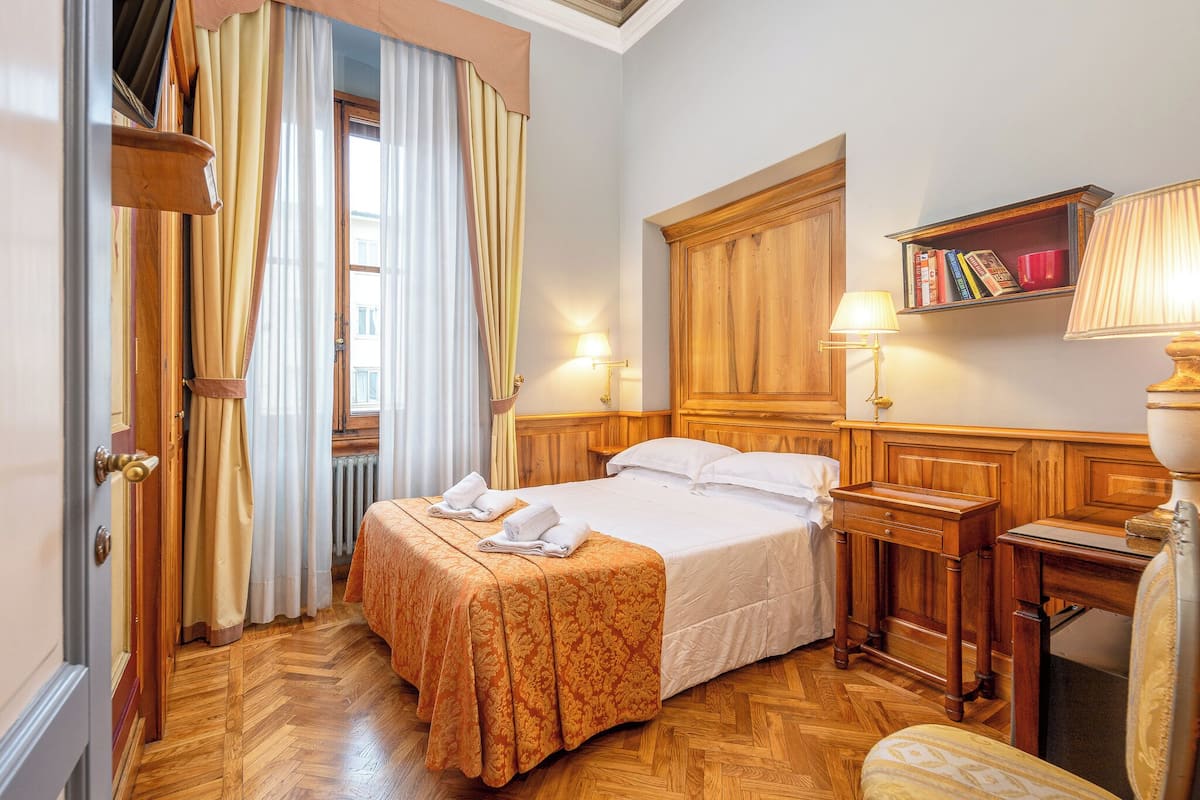 A hotel room with a bed, wooden headboard, bedside tables, a lamp, a bookshelf, and a window with curtains.