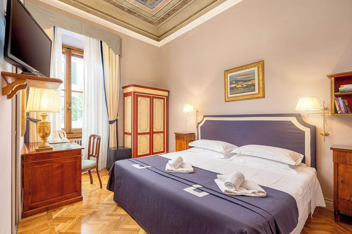 A hotel room with a large bed, a television, a desk with a lamp, a wardrobe, and a painting on the wall.