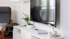 A modern living room with a flat-screen TV mounted on a white wall, a grey armchair, a white console table with a vase of flowers, and a remote control. - Marci's Home (Rome)