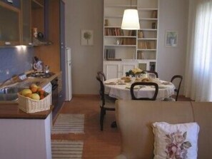 A cozy dining area with a table set for two, a bookshelf, and a kitchen area with a fruit basket. - Marci's Home (Rome)