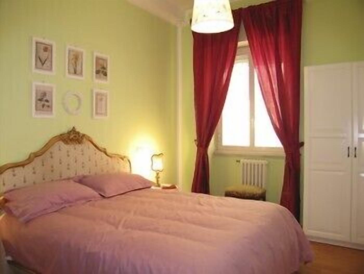 A bedroom with a bed, a nightstand, a window with red curtains, and wall-mounted pictures.