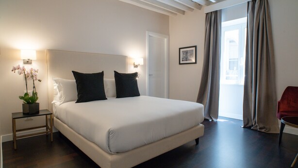 A bedroom with a large bed, a nightstand with a lamp, a potted plant, and a red chair. - Kame Hall (Rome)