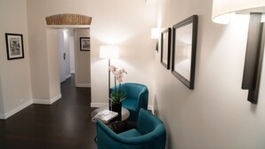 A hallway with dark wooden flooring, a teal armchair, a small table with a magazine, a potted orchid, and framed pictures on the wall. - Kame Hall (Rome)