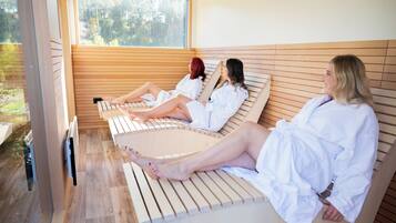 Three individuals relaxing on wooden loungers in a room with large windows and a view of a forest.