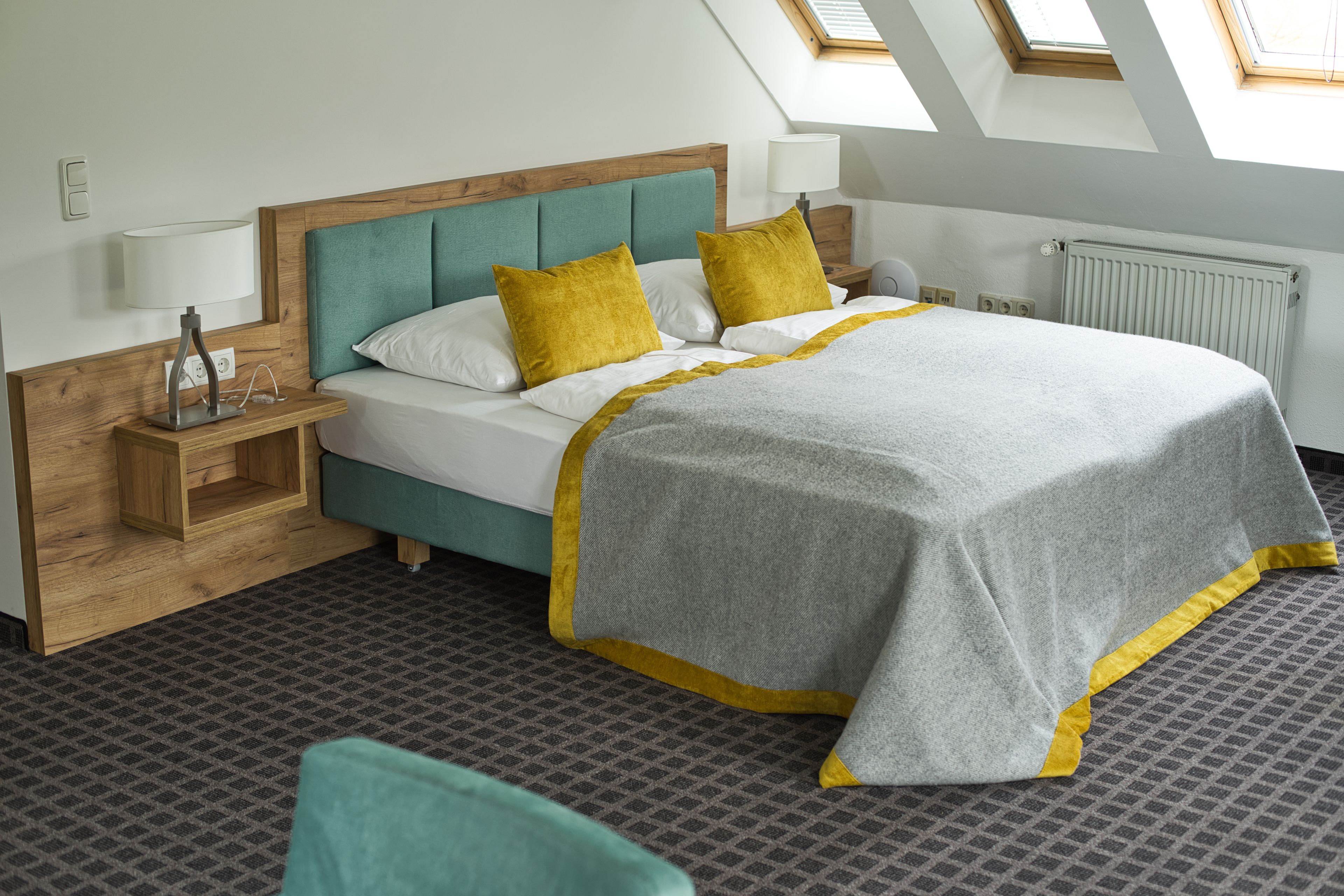 Une chambre d’hôtel bien rangée, avec un lit, une table de chevet, une lampe et une fenêtre avec des lucarnes.