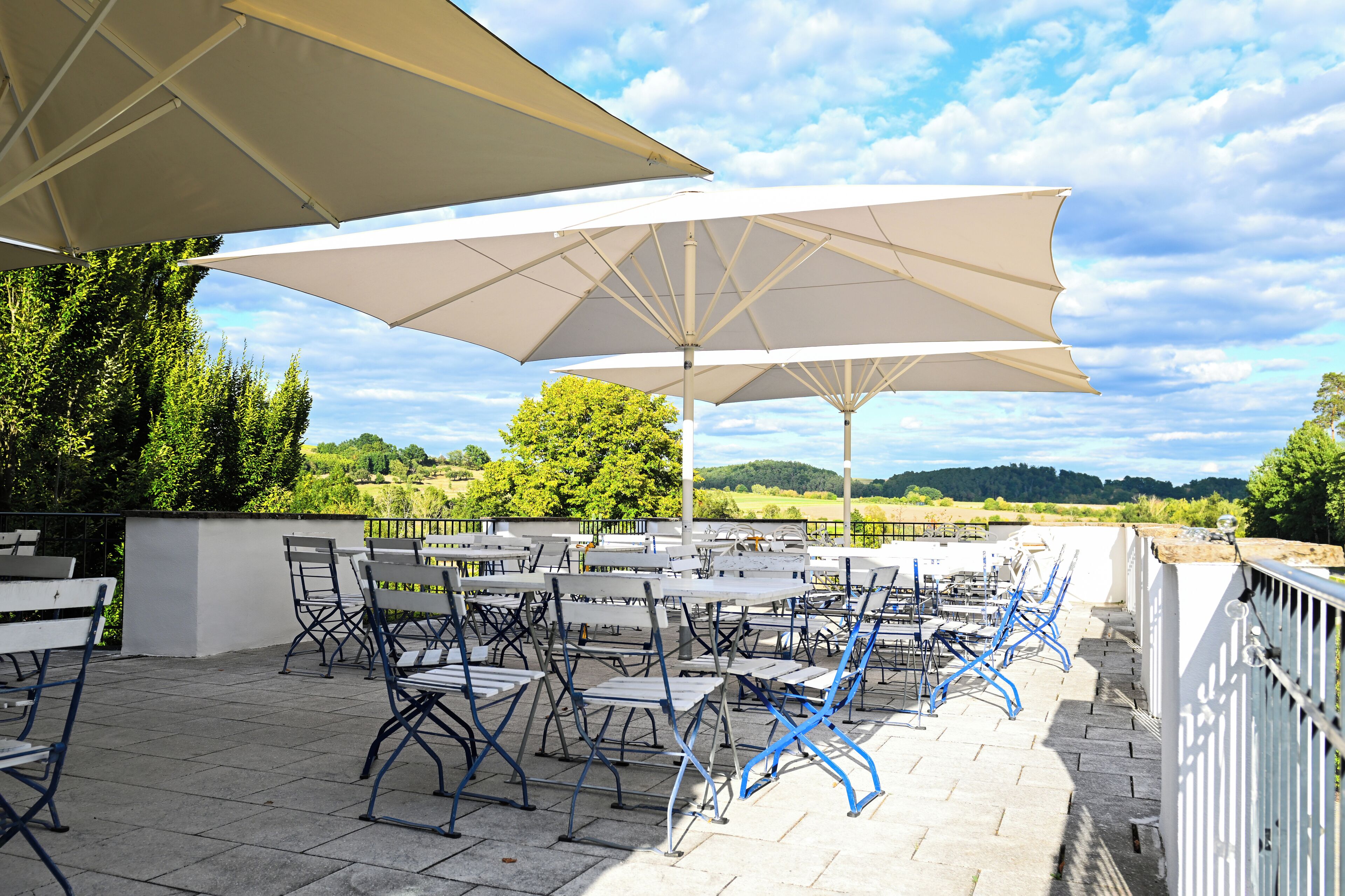 Een terras buiten met witte tafels en blauwe stoelen, grote witte parasols en een mooi uitzicht op heuvels en bomen.