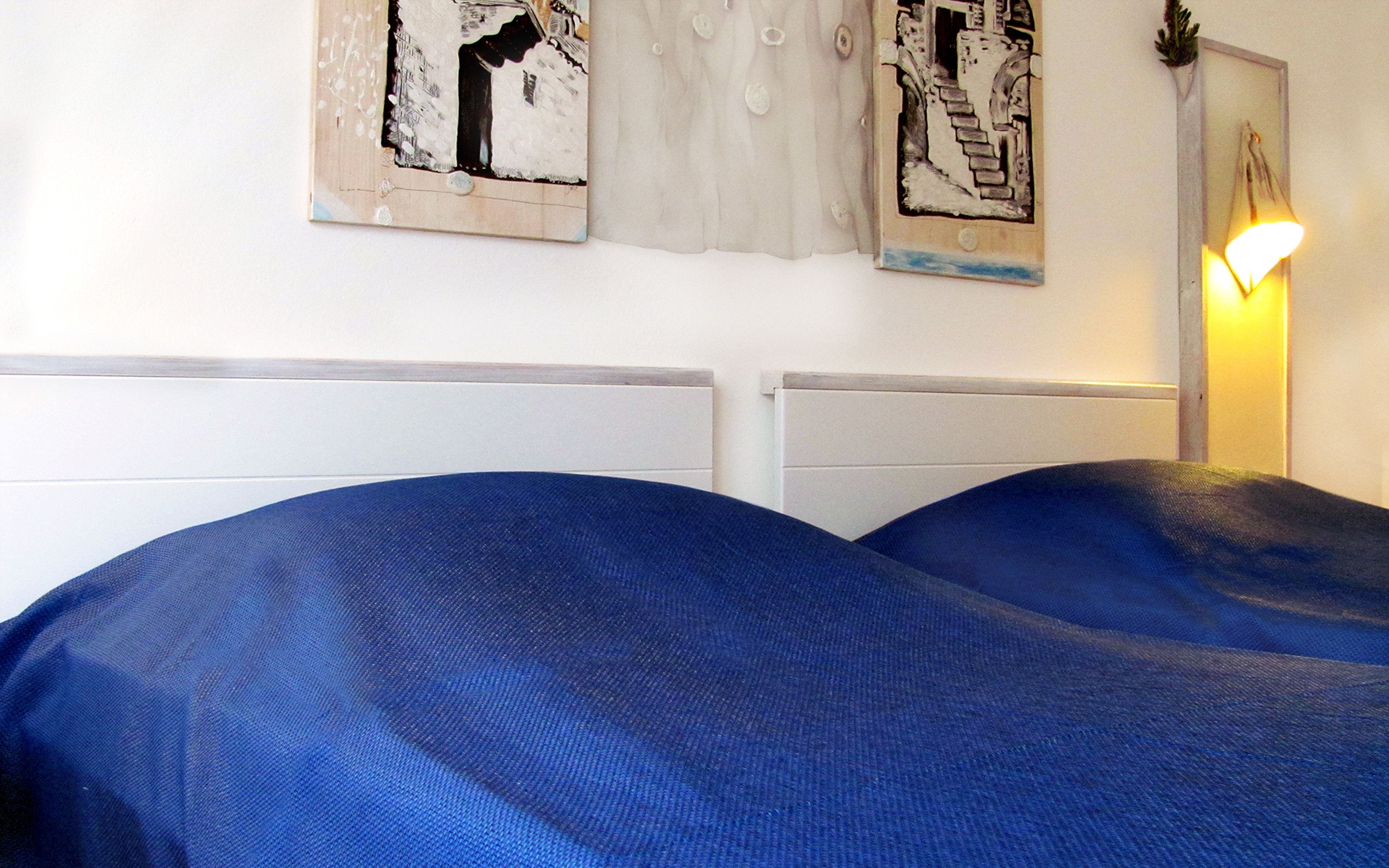Une chambre avec deux lits recouverts de draps bleus, des têtes de lit blanches et un luminaire fixé au mur.