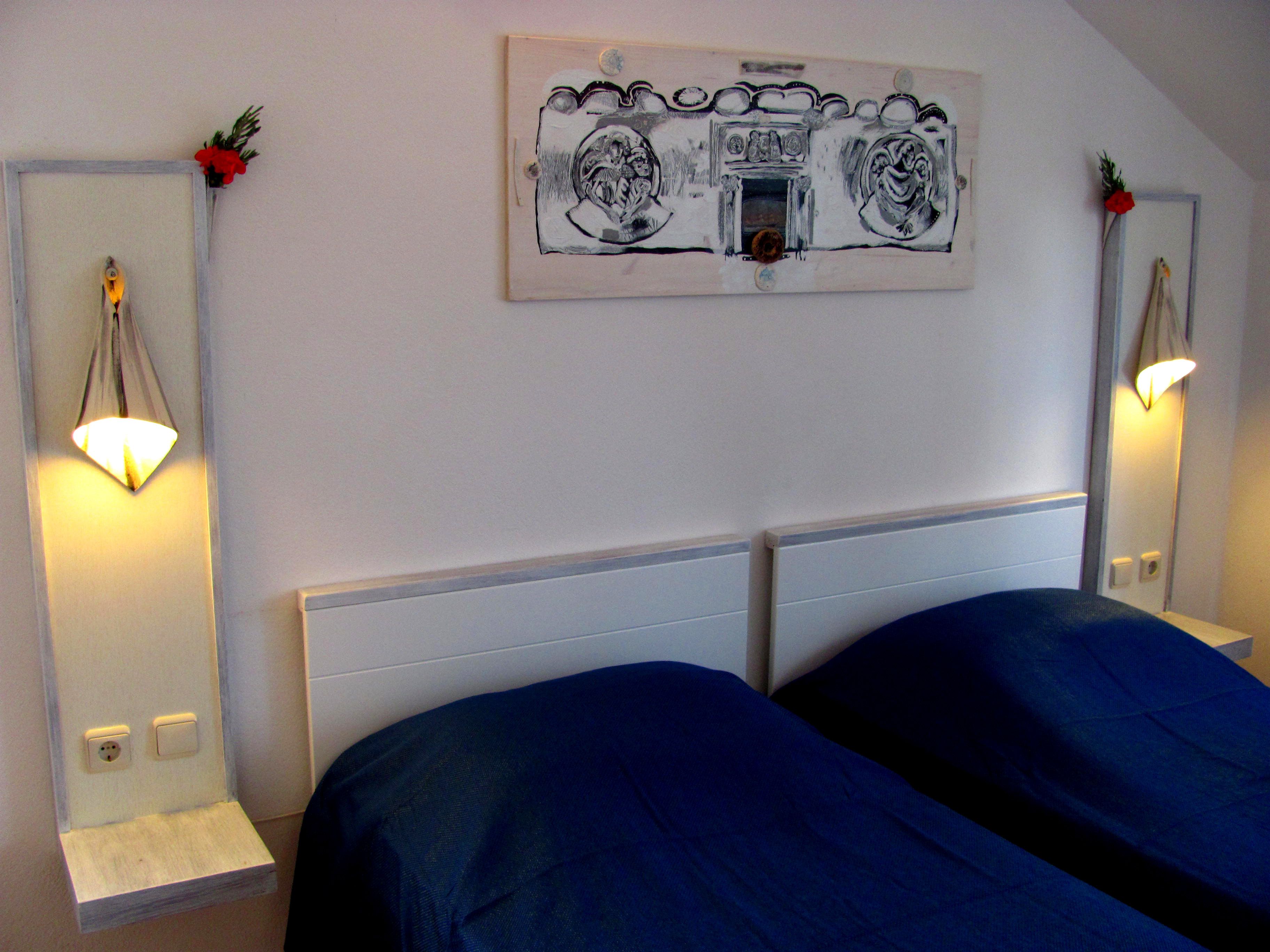 A bedroom with a double bed, two bedside lamps, a framed artwork above the bed, and a small shelf with a plant.