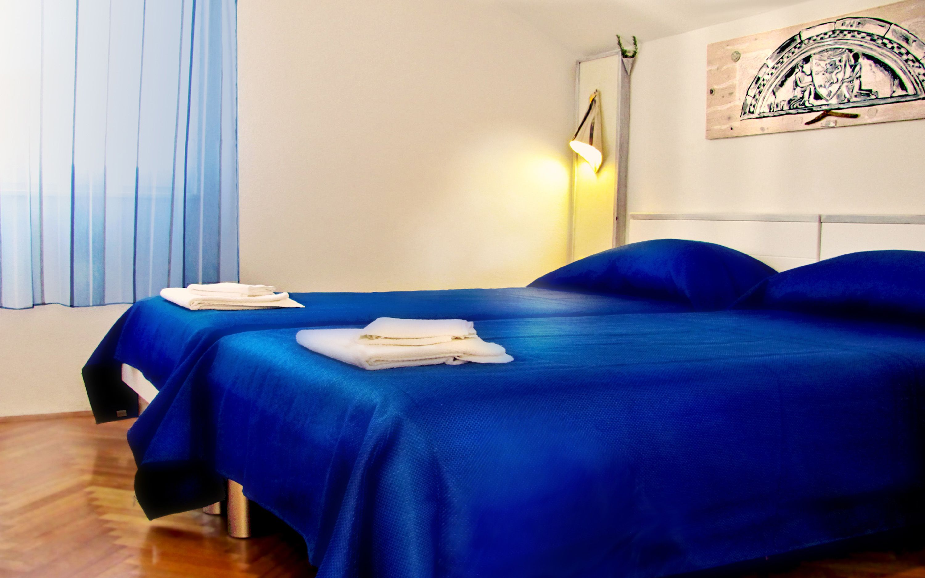 A hotel room with two beds, a wooden floor, a wall-mounted lamp, and a painting.