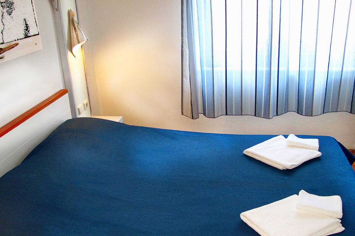 Una habitación de hotel con una colcha azul, dos toallas blancas dobladas con cuidado, una ventana con persianas verticales y una lámpara fijada a la pared.