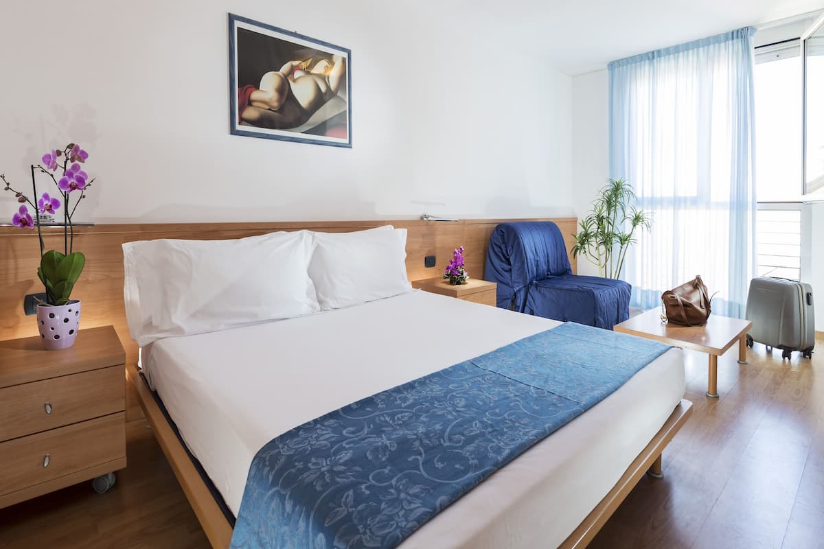 A hotel room with a large bed, a wooden nightstand, a desk, a chair, a suitcase, and a potted plant.