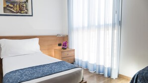 Un lit bien fait, recouvert d’une couvre-lit bleue à motifs, une table de chevet en bois ornée d’un vase de fleurs violettes et de fines rideaux laissant passer la lumière naturelle.