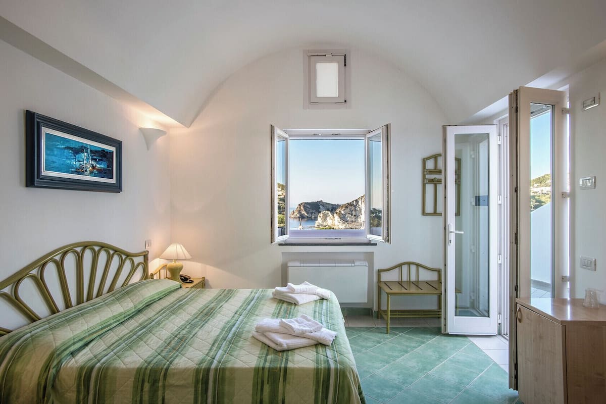 Une chambre à coucher avec un lit, un bureau placé près d’une fenêtre donnant sur des falaises, une chaise et une porte menant à une autre pièce.
