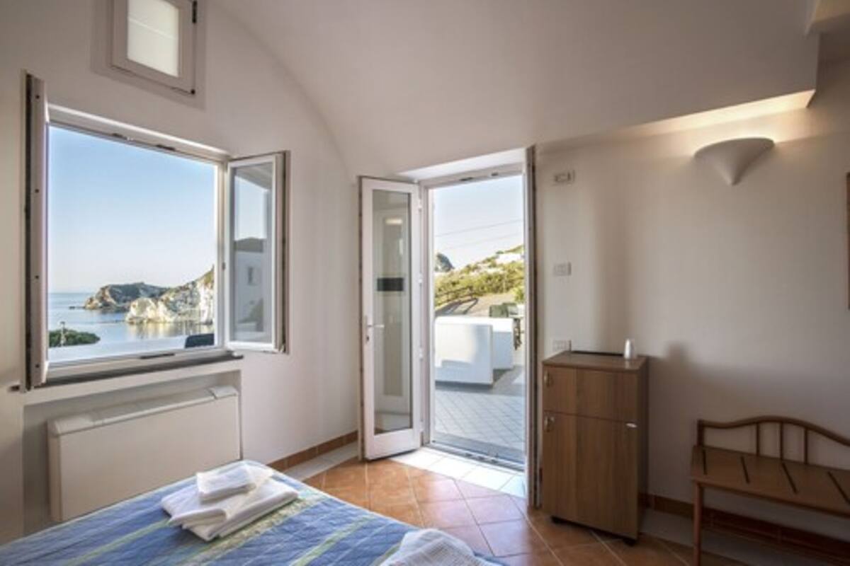 Une chambre à coucher avec un lit, une armoire en bois et une vue sur la mer, visible à travers une grande fenêtre.
