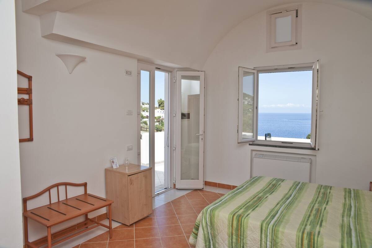 Une chambre à coucher comprenant un lit, un banc en bois, une petite armoire et une porte-fenêtre coulissante donnant sur un balcon avec vue sur la mer.