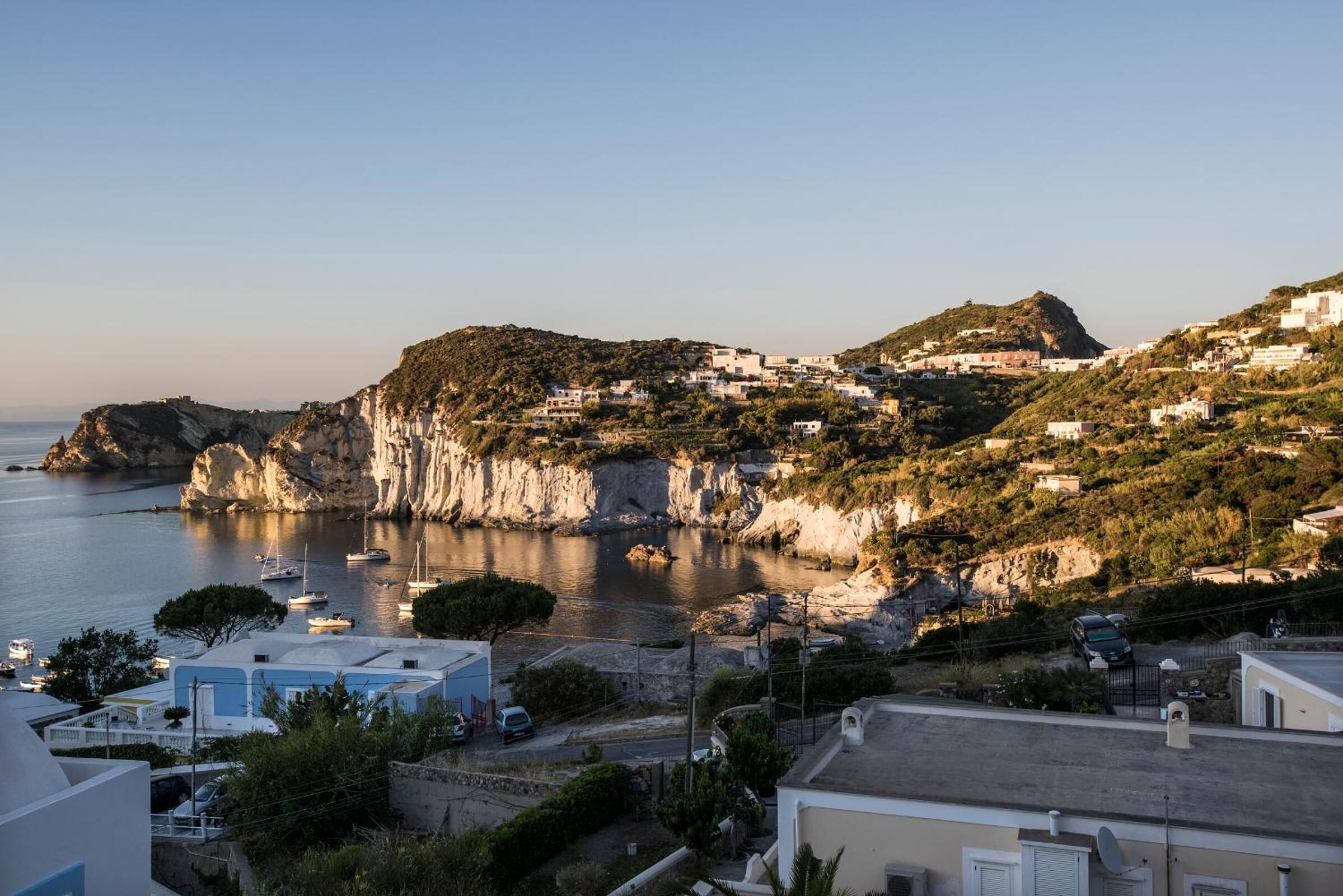 A coastal town with buildings on a hillside overlooking the sea, a rocky coastline, and boats anchored near the shore.