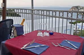 A table set for two with a red tablecloth, two wine glasses, a bottle of champagne, and a view of the sea and mountains in the background.