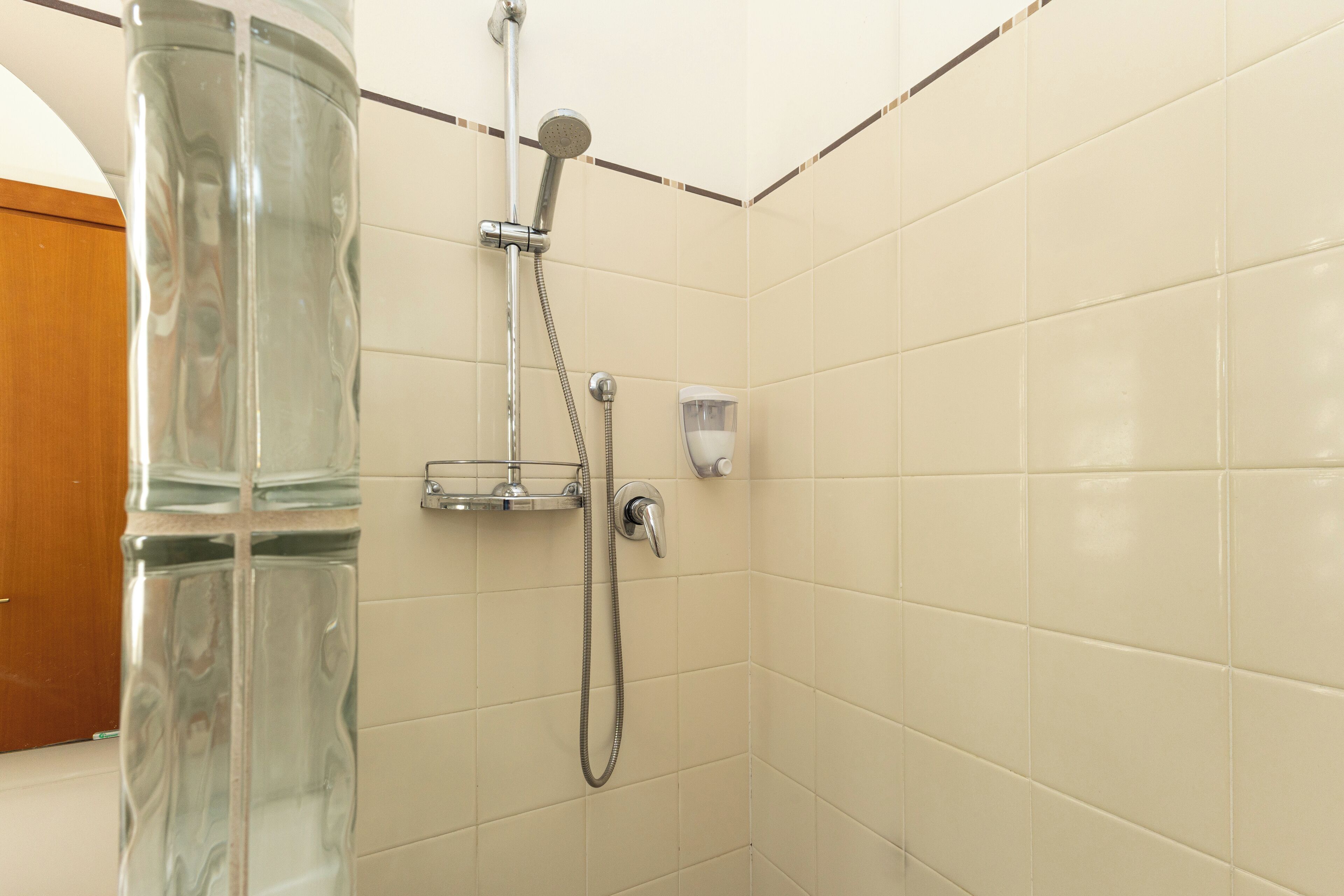 Ducha con mampara de vidrio, cabezal de ducha de mano y dispensador de jabón en la pared.