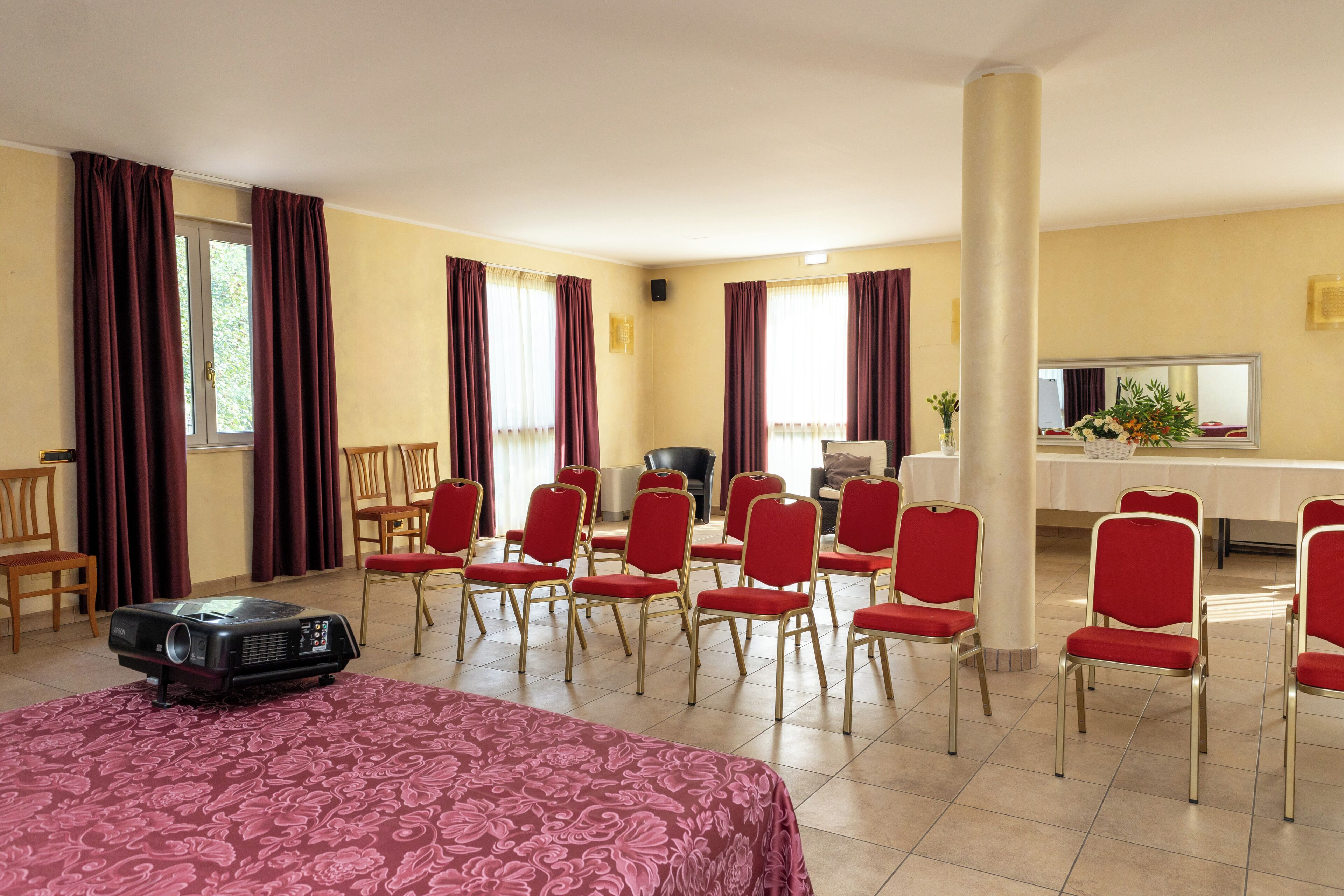 Una habitación de hotel con una cama, un proyector, sillas rojas, un escritorio y un pilar grande.