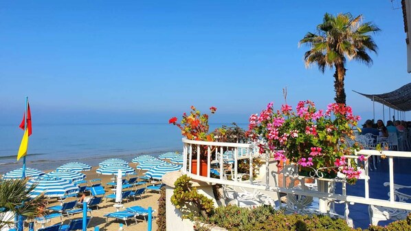 Un hôtel situé en bord de mer, doté de parasols à rayures bleues, de chaises longues et offrant une vue sur l’océan.