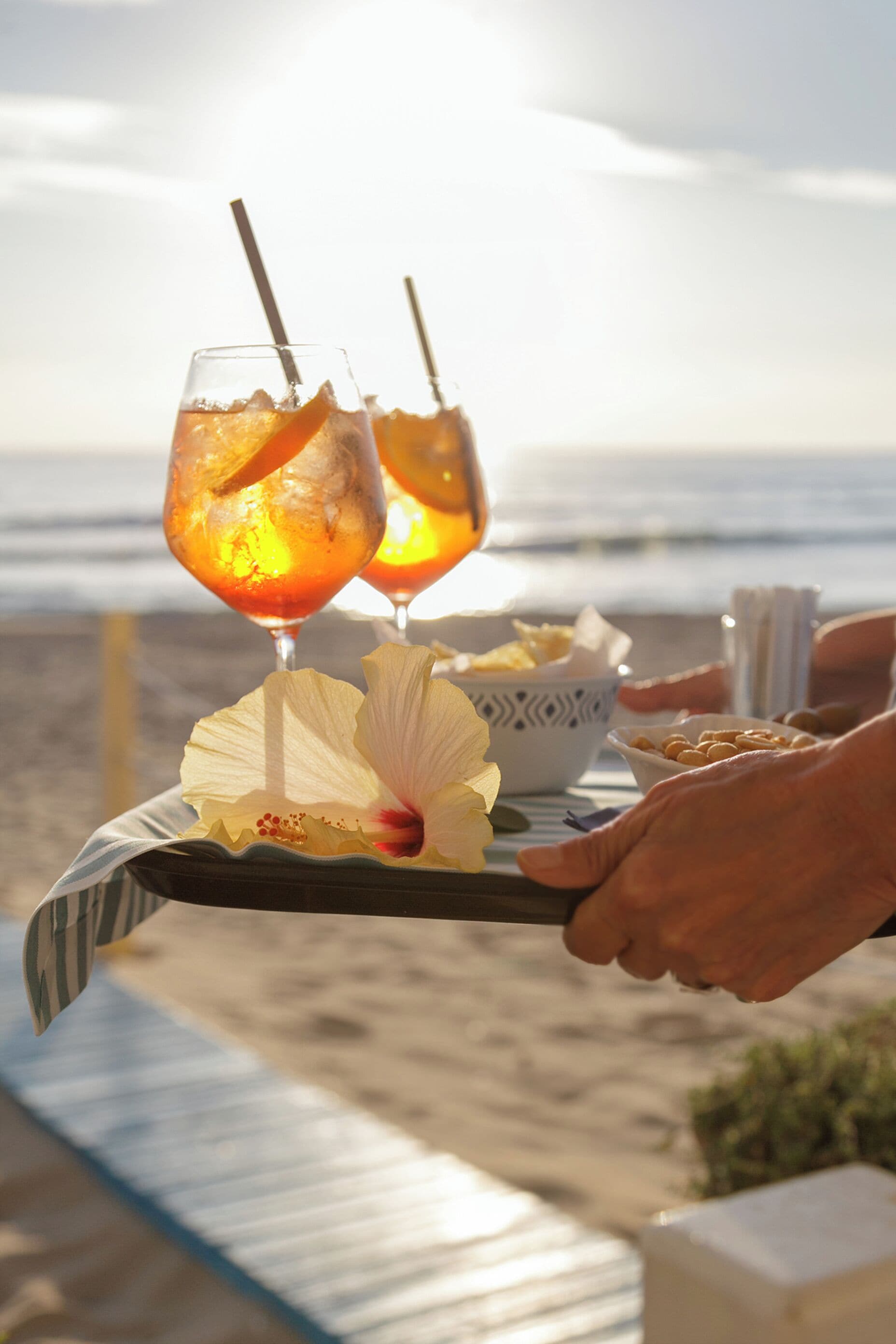 A tray with two glasses of a beverage, a flower, and a bowl of snacks on a beach.