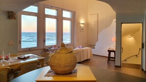 A room with a large arched window overlooking the sea, a bed with a white bedspread, a table with a woven basket, and a lamp on a side table.