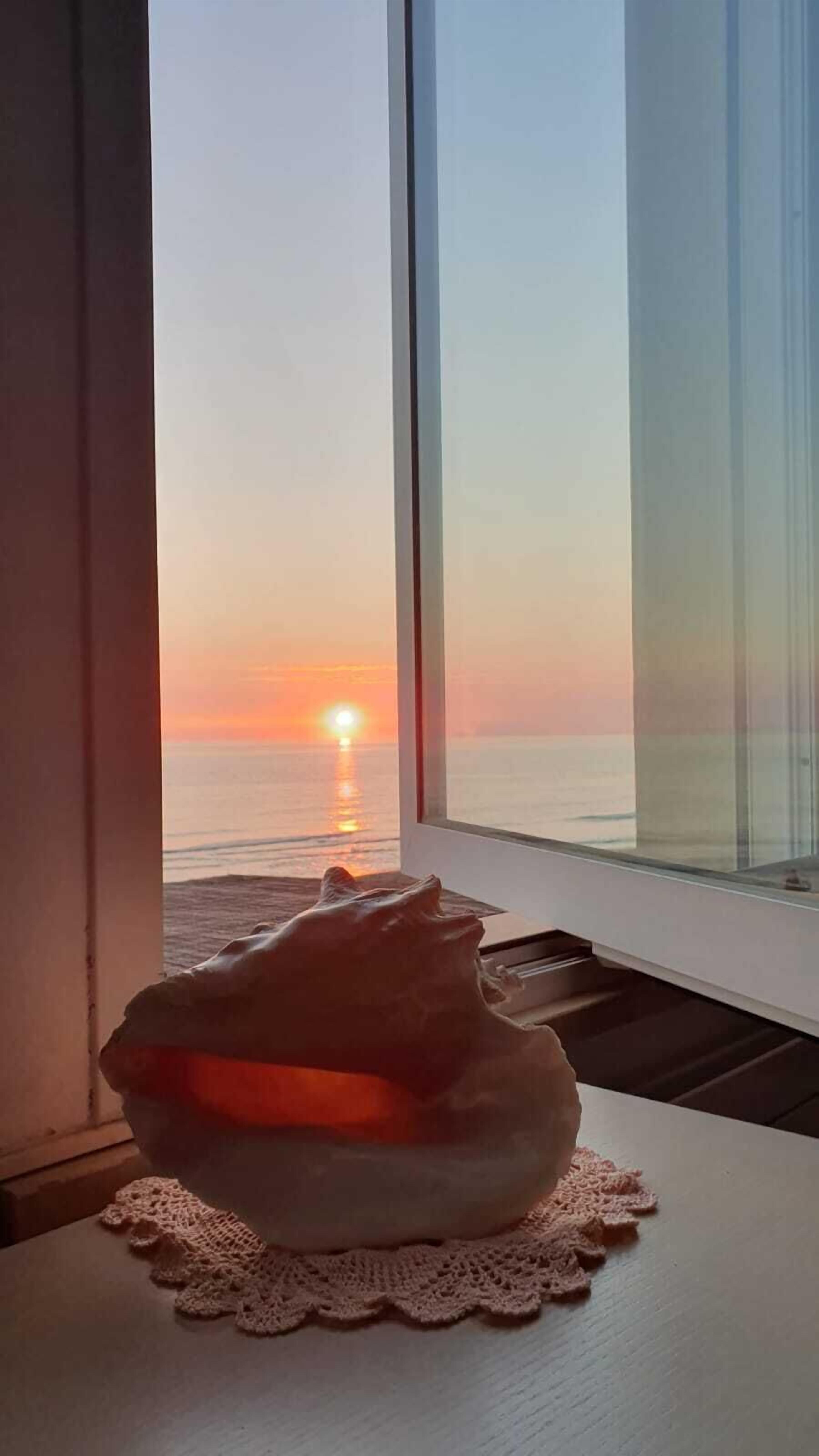Vista del océano al atardecer a través de una ventana, con una vela con forma de gato sobre un posavasos de encaje.