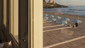 A beach view from a room with large windows, featuring a wooden deck with beach umbrellas and a castle-like structure on the shore.