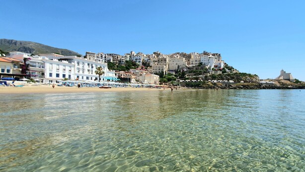 A coastal town with clear waters, buildings on a hillside, and a fort on a small island.