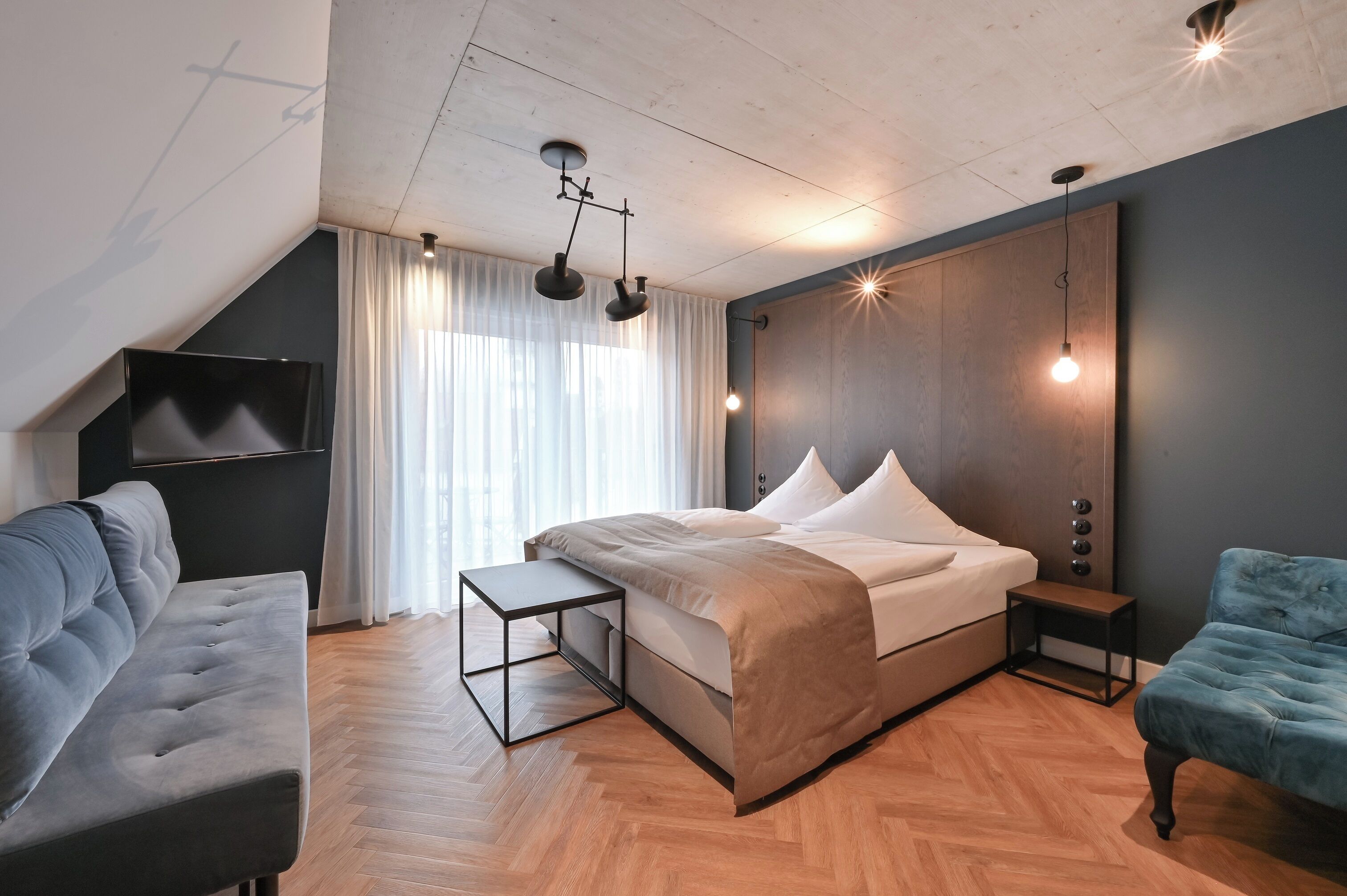 Une chambre à coucher moderne avec un grand lit, un canapé gris, une petite table et un fauteuil bleu.