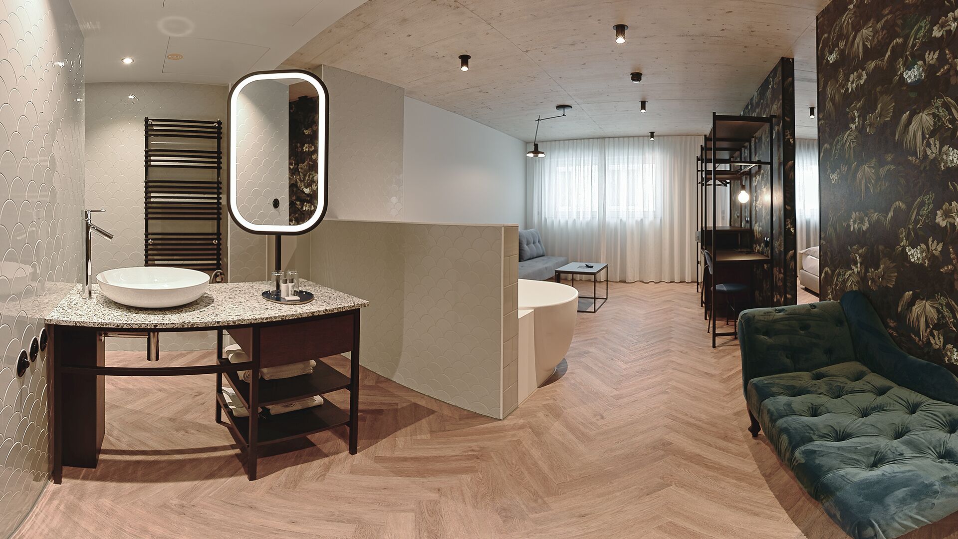 A modern bathroom with a vanity, sink, and mirror. There is a wooden herringbone floor and a green upholstered sofa in the living area.