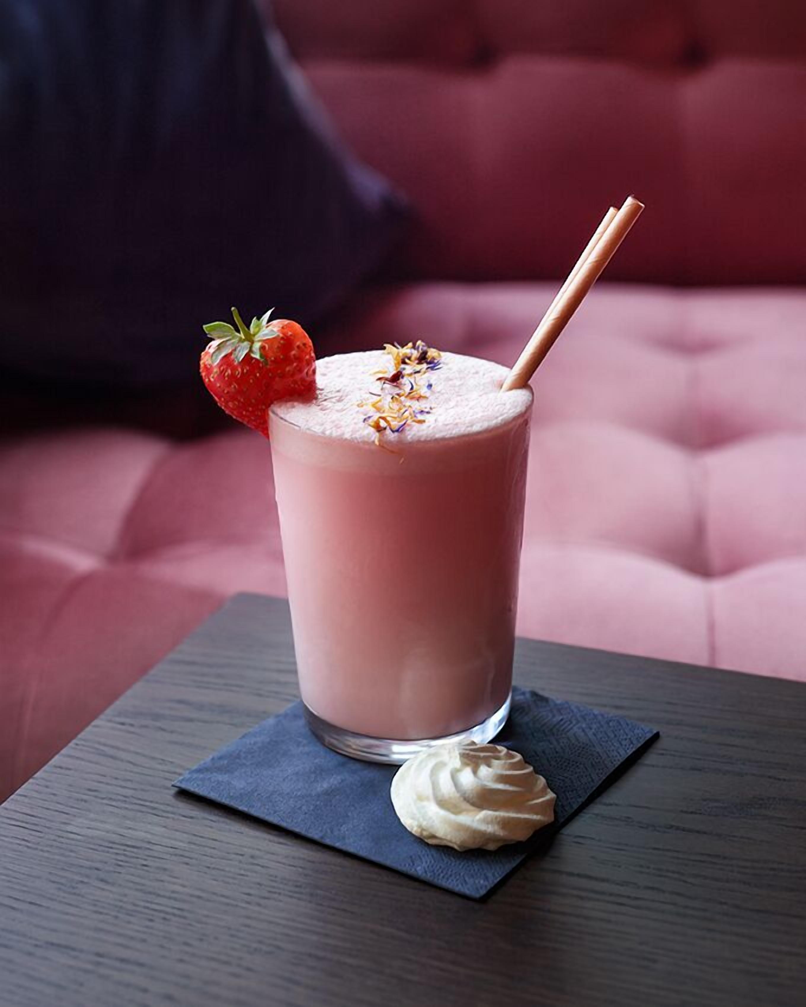 Una bebida rosada con una decoración de fresa y una cobertura de crema batida sobre un posavasos oscuro, colocada sobre una mesa con un sofá rojo acolchado al fondo.