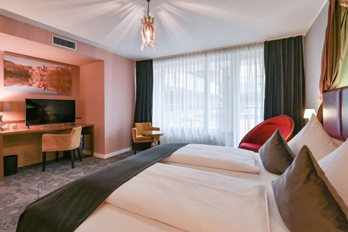 Una habitación de hotel con cama, escritorio, televisor y un gran ventanal con cortinas.