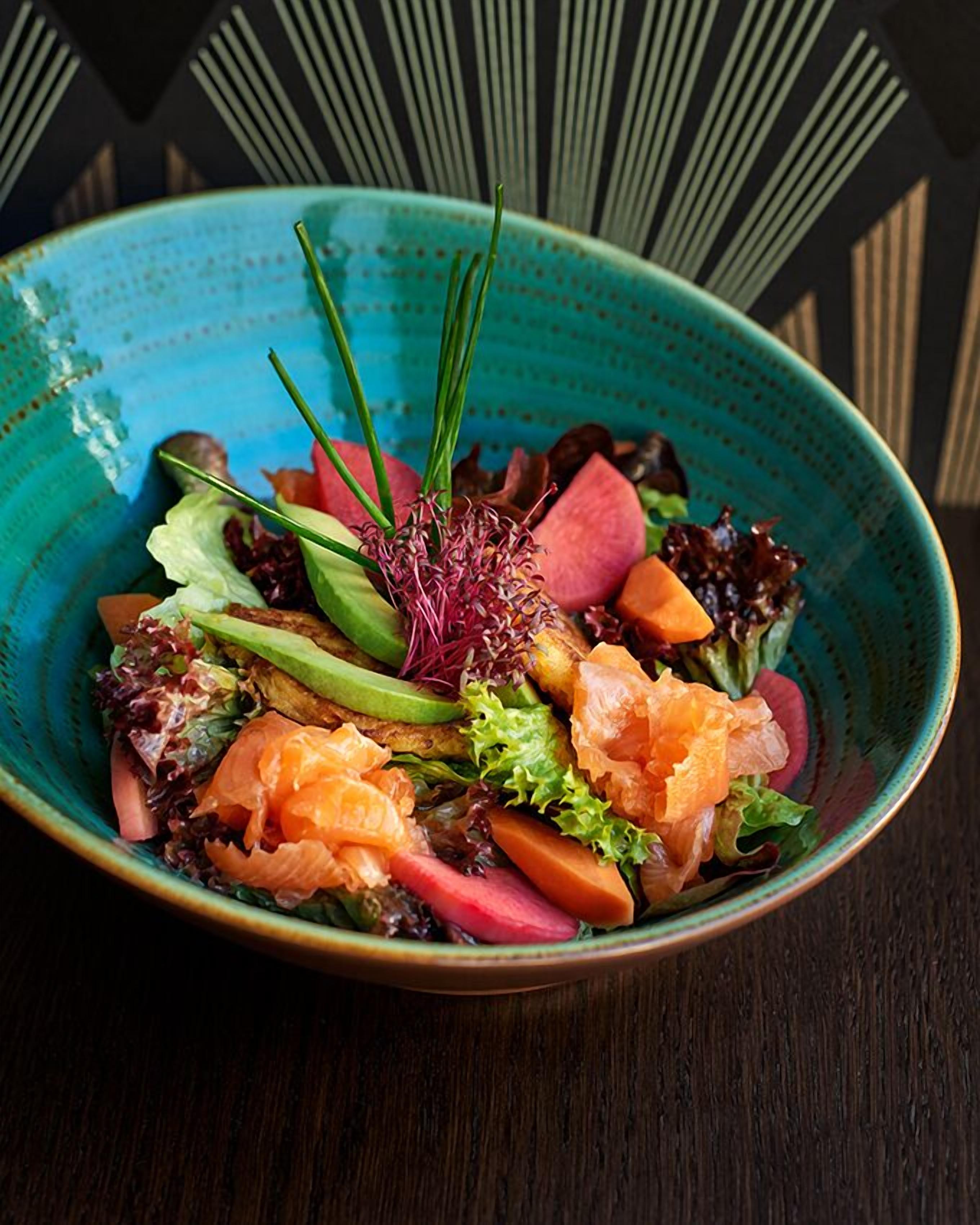 Un tazón de ensalada con varias verduras y salmón, sobre una mesa de madera oscura.