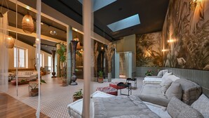 Habitación amplia con una cama grande, un sofá y una zona de comedor. Hay plantas y un mural en la pared.