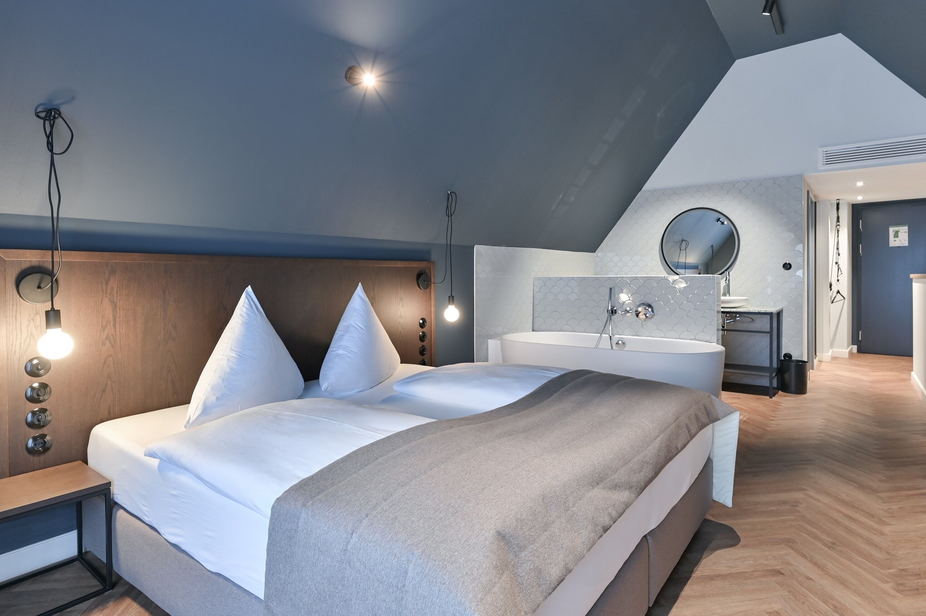 Un dormitorio moderno con una cama grande, una mesita de noche, un banco empotrado, un espejo redondo y un baño con bañera y lavamanos.