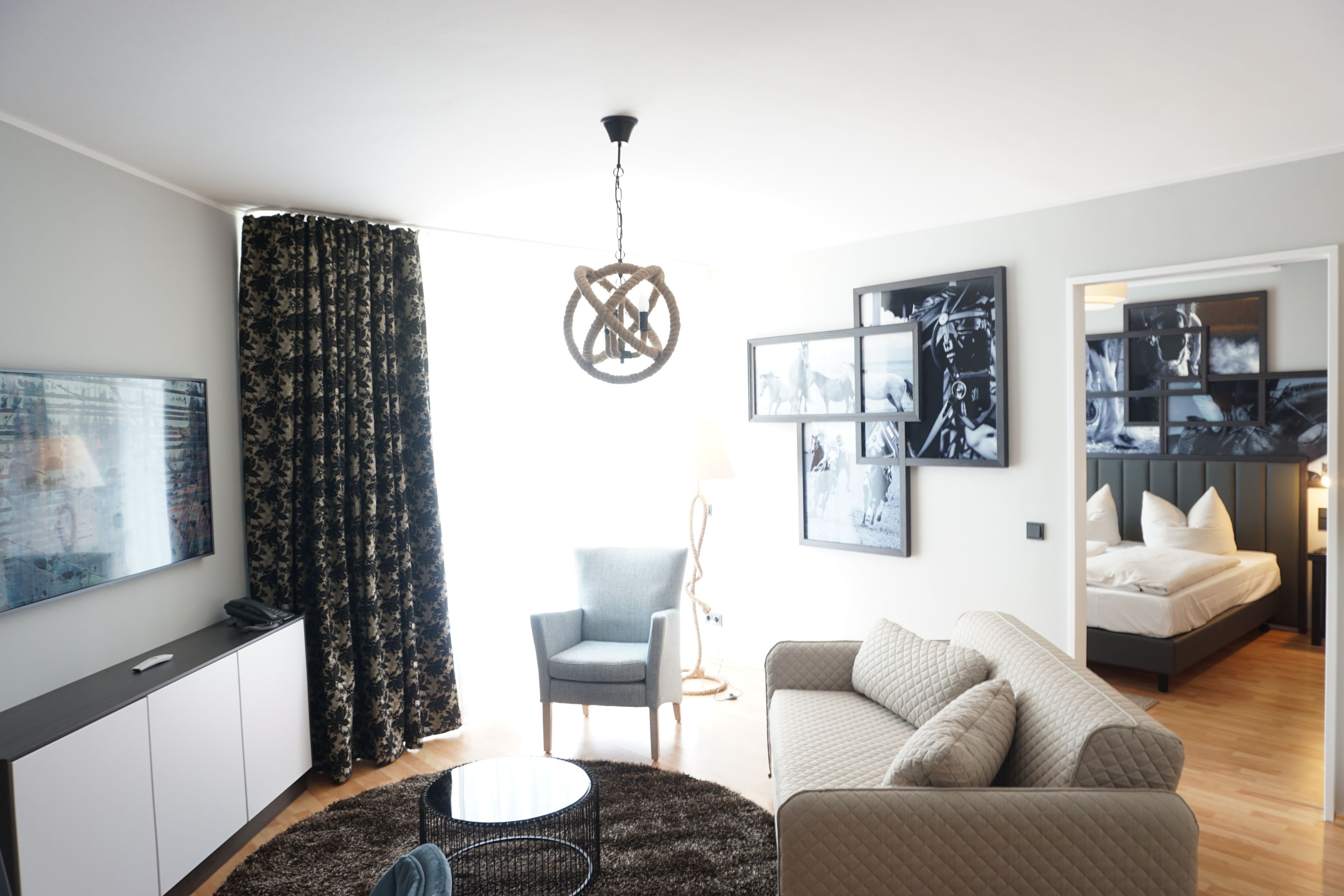 Una sala de estar moderna con un sofá, un sillón y una mesa de centro redonda. Se ve una cama con cabecera al fondo, y obras de arte en las paredes.