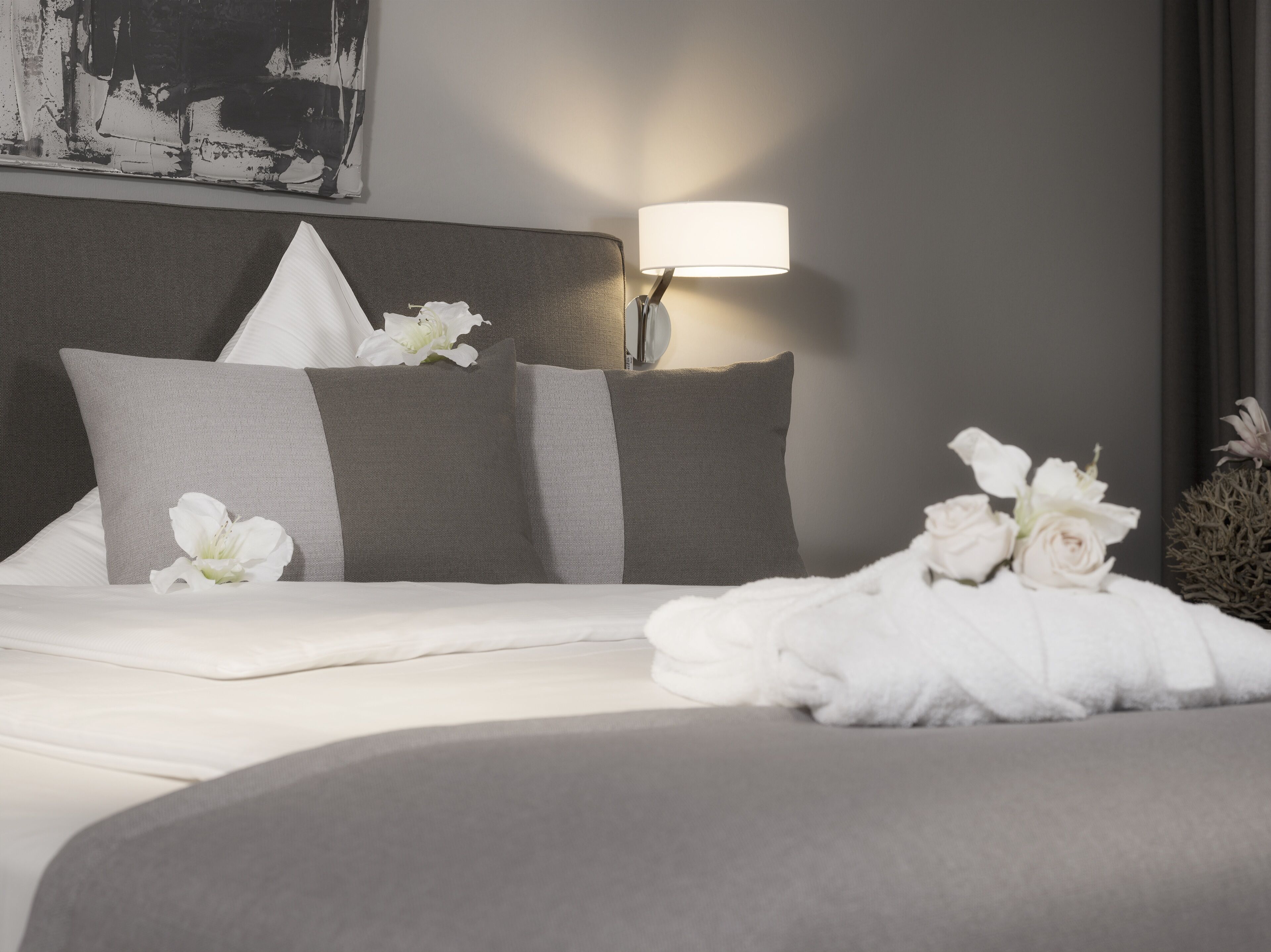 A neatly made bed with white linens and gray pillows, a bedside lamp, and a bouquet of white flowers on the bed.