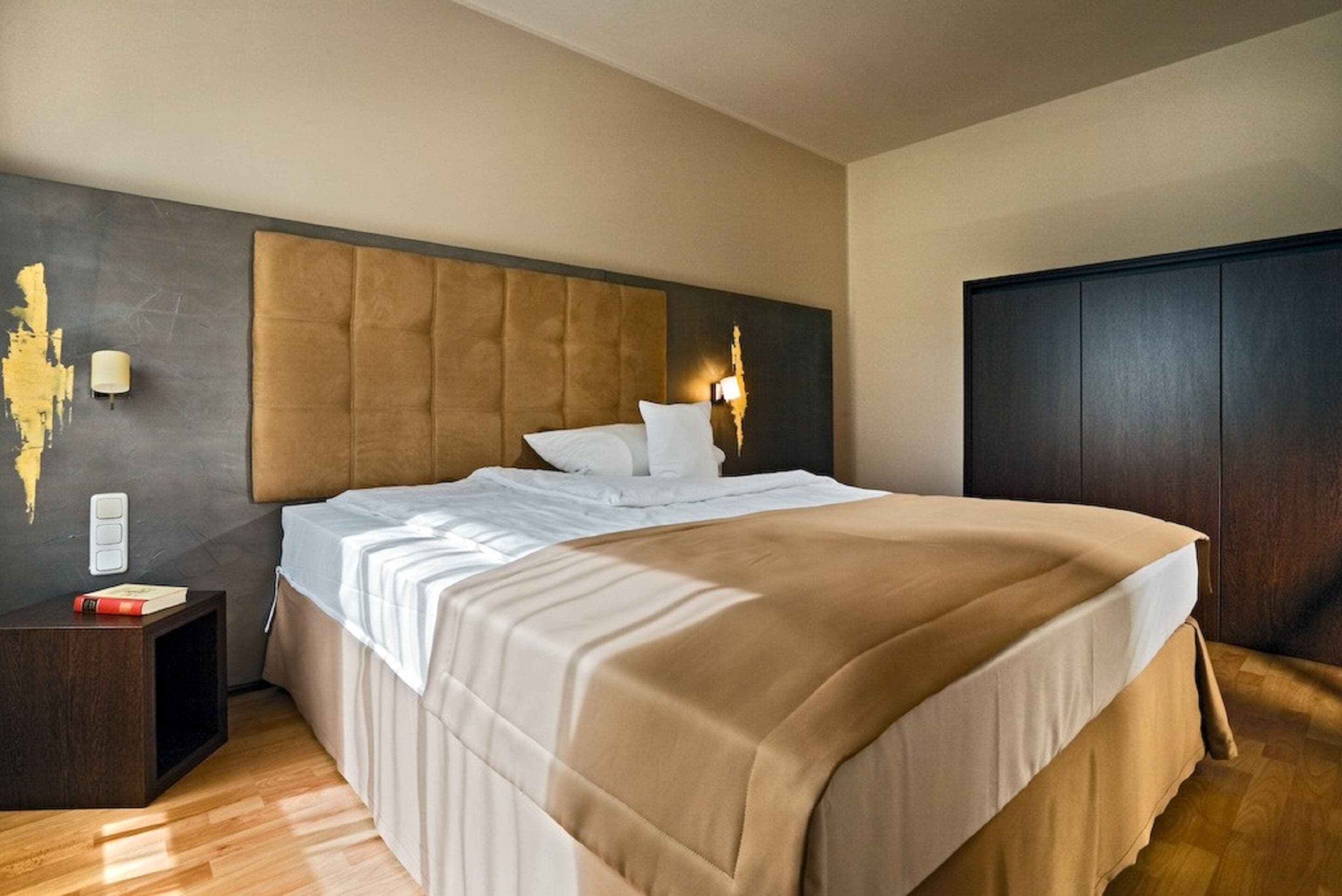 A modern hotel room with a large bed, a headboard with a light fixture, a nightstand, and a dark wooden wardrobe.