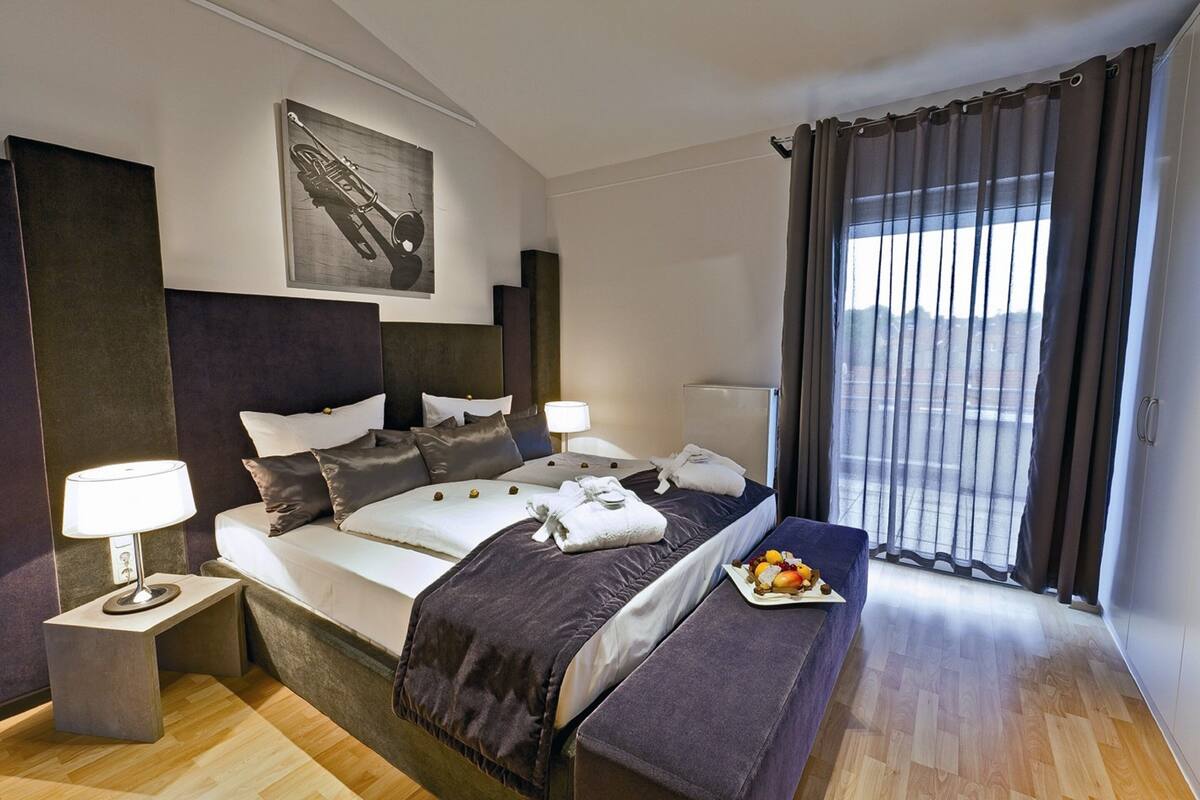 Une chambre d’hôtel moderne avec un grand lit, des tables de chevet, un banc et une vue sur l’extérieur.