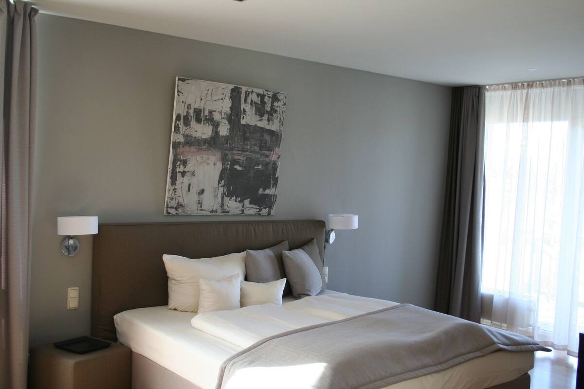 Une chambre d’hôtel avec un grand lit, deux lampes de chevet, une œuvre d’art accrochée au mur et une fenêtre avec des rideaux.