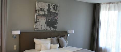 Habitación de hotel con una cama grande, dos lámparas de noche, un cuadro en la pared y una ventana con cortinas.