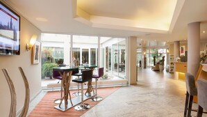 A spacious hotel lobby with a modern design, featuring a glass partition, a reception desk, and a dining area with a glass table and chairs.