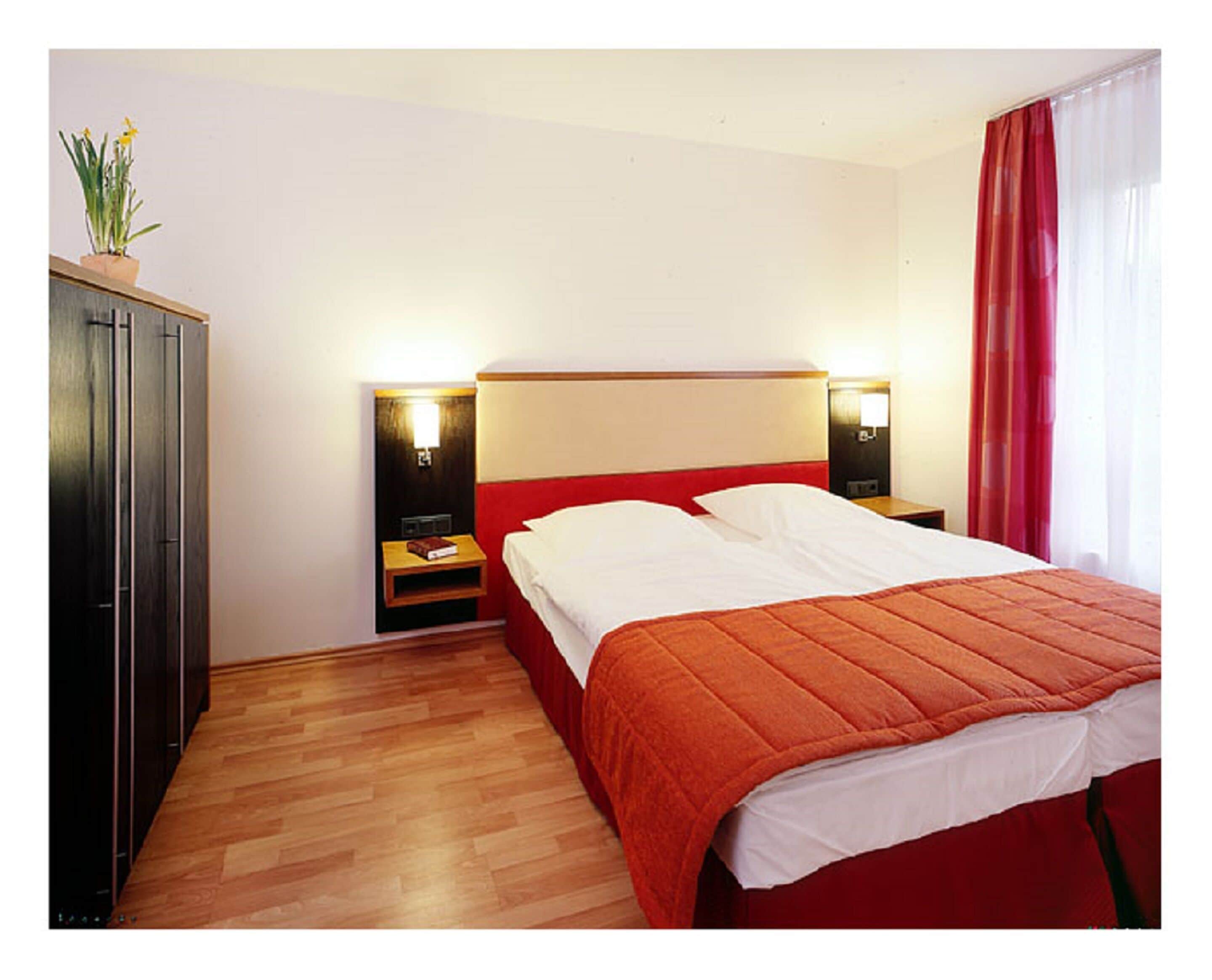 Habitación de hotel con piso de madera, cama con cabecera roja, dos mesitas de noche con lámparas, armario y ventana con cortinas rojas.