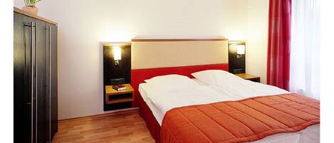 Habitación de hotel con piso de madera, cama con cabecera roja, dos mesitas de noche con lámparas, armario y ventana con cortinas rojas.
