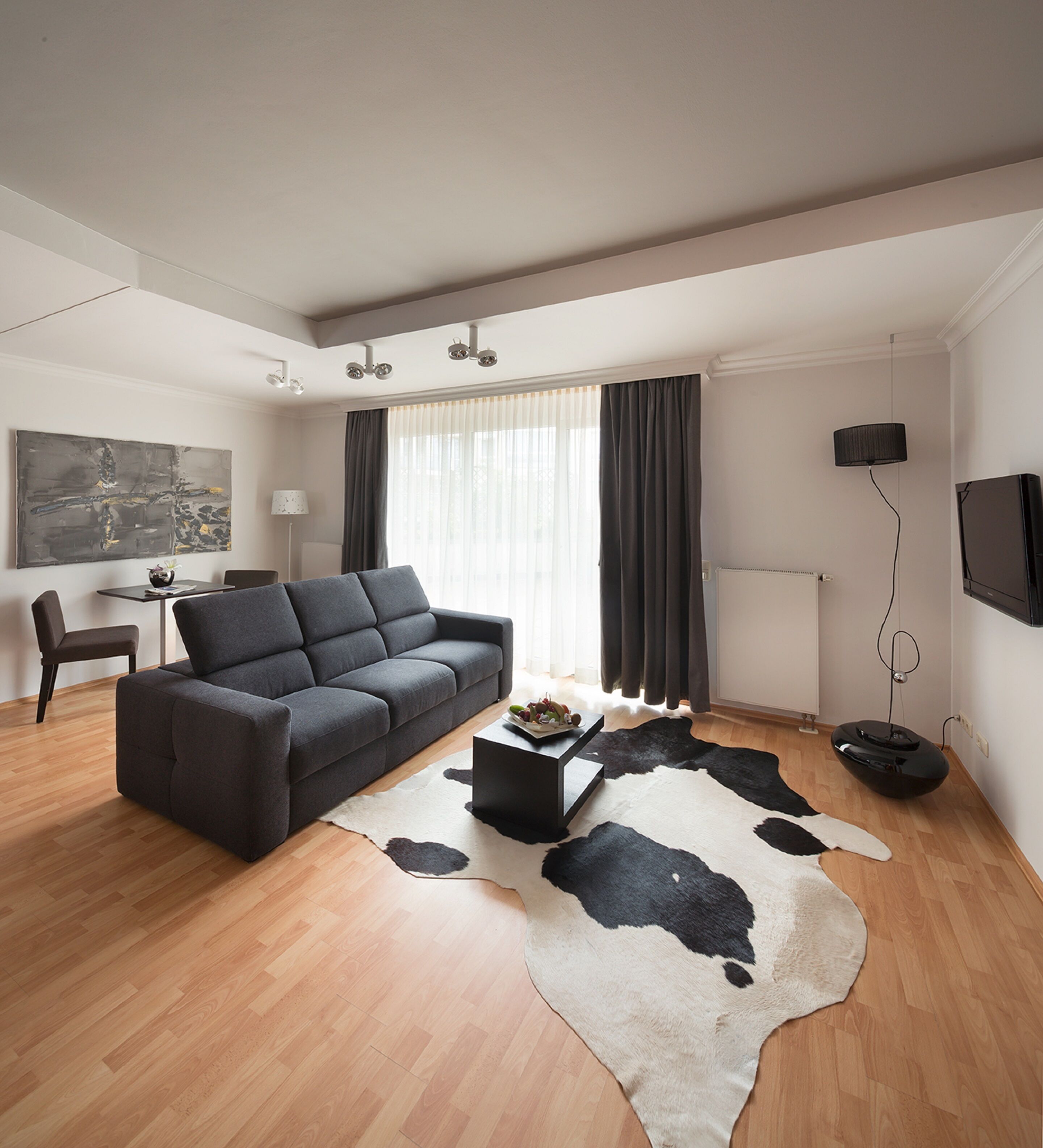 Un salon moderne avec un canapé gris, une table basse ronde, un tapis en peau de vache noir et blanc, un coin repas avec une table et des chaises, et un téléviseur fixé au mur.
