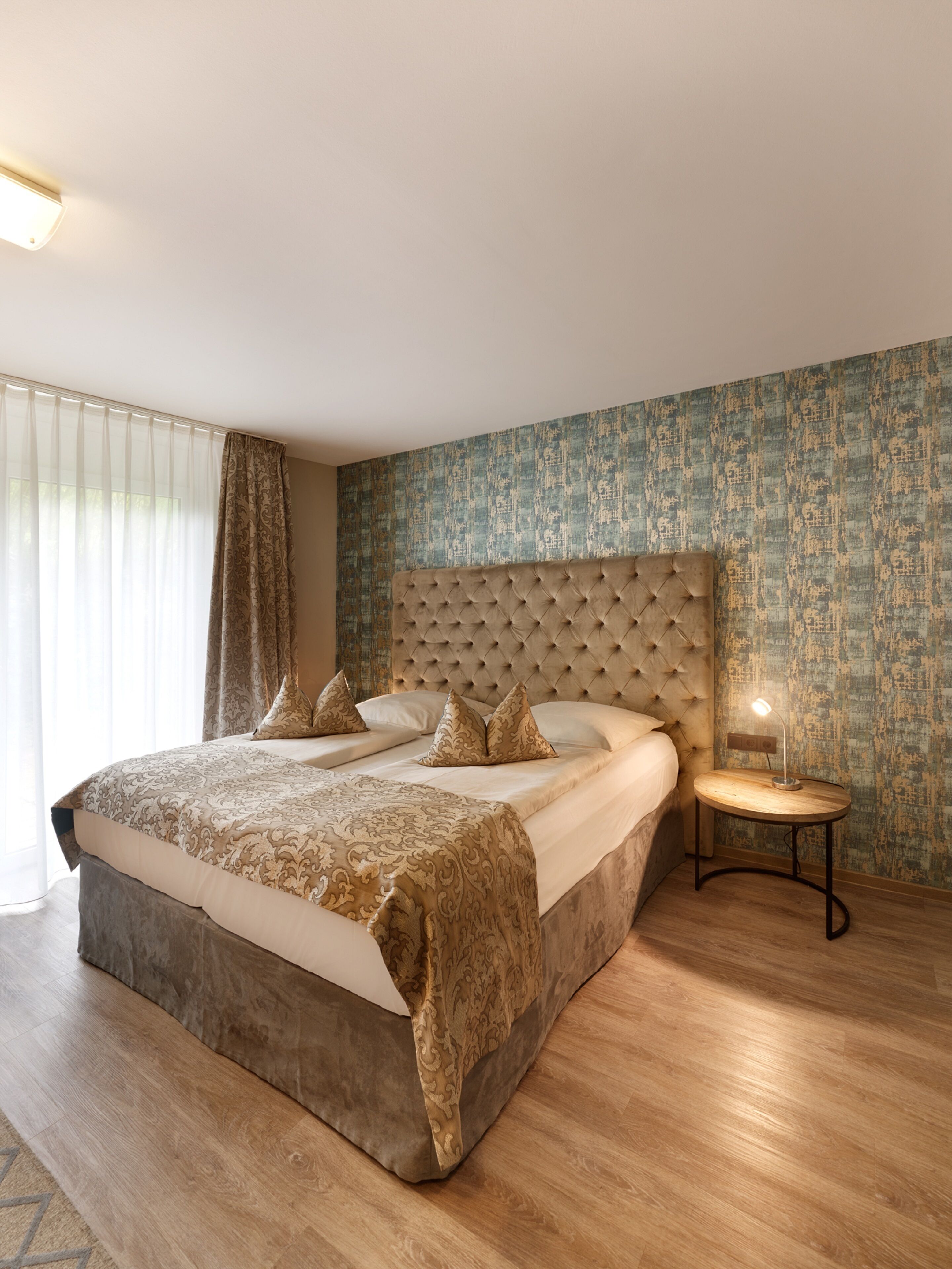 A large bed with a tufted headboard, a bedside table with a lamp, and a patterned wall.